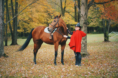 rider on ground next to bay mare. Rider wears a red cape and witches hat.